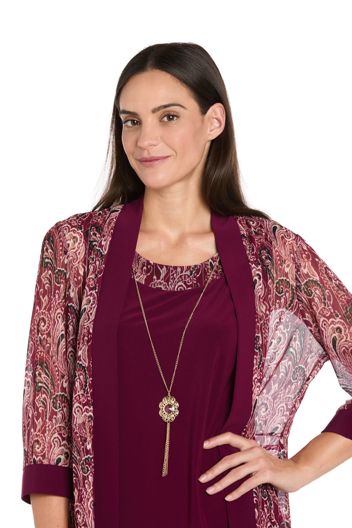 A woman with long brown hair confidently poses in the R&M Richards 3029P Petite Short Wine Party Dress, perfect for semi-formal occasions. She accessorizes with a gold pendant necklace over a maroon top.